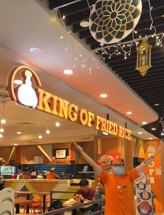 A King of Fried Rice crew member in the orange house tee under the lit King of Fried Rice signboard, with diners seated behind